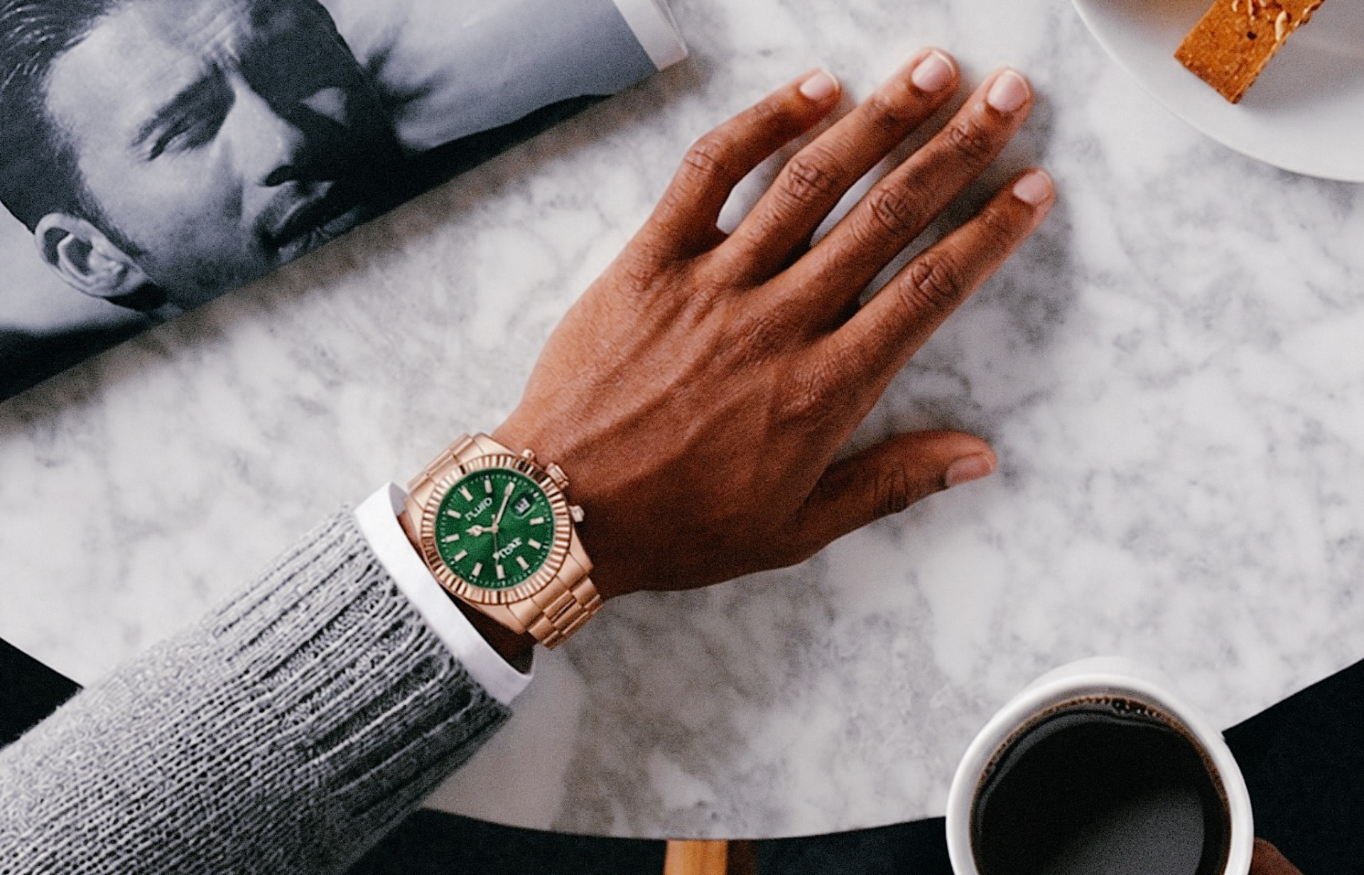 Green-dial gold watch on a marble table beside a coffee and a magazine